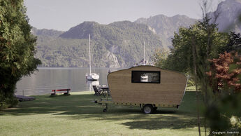 Der kleine Holz-Wohnwagen steht am Ufer eines Sees. Im Hintergrund segelt ein Boot vorbei und bewaldete Hügel säumen das Wasser.